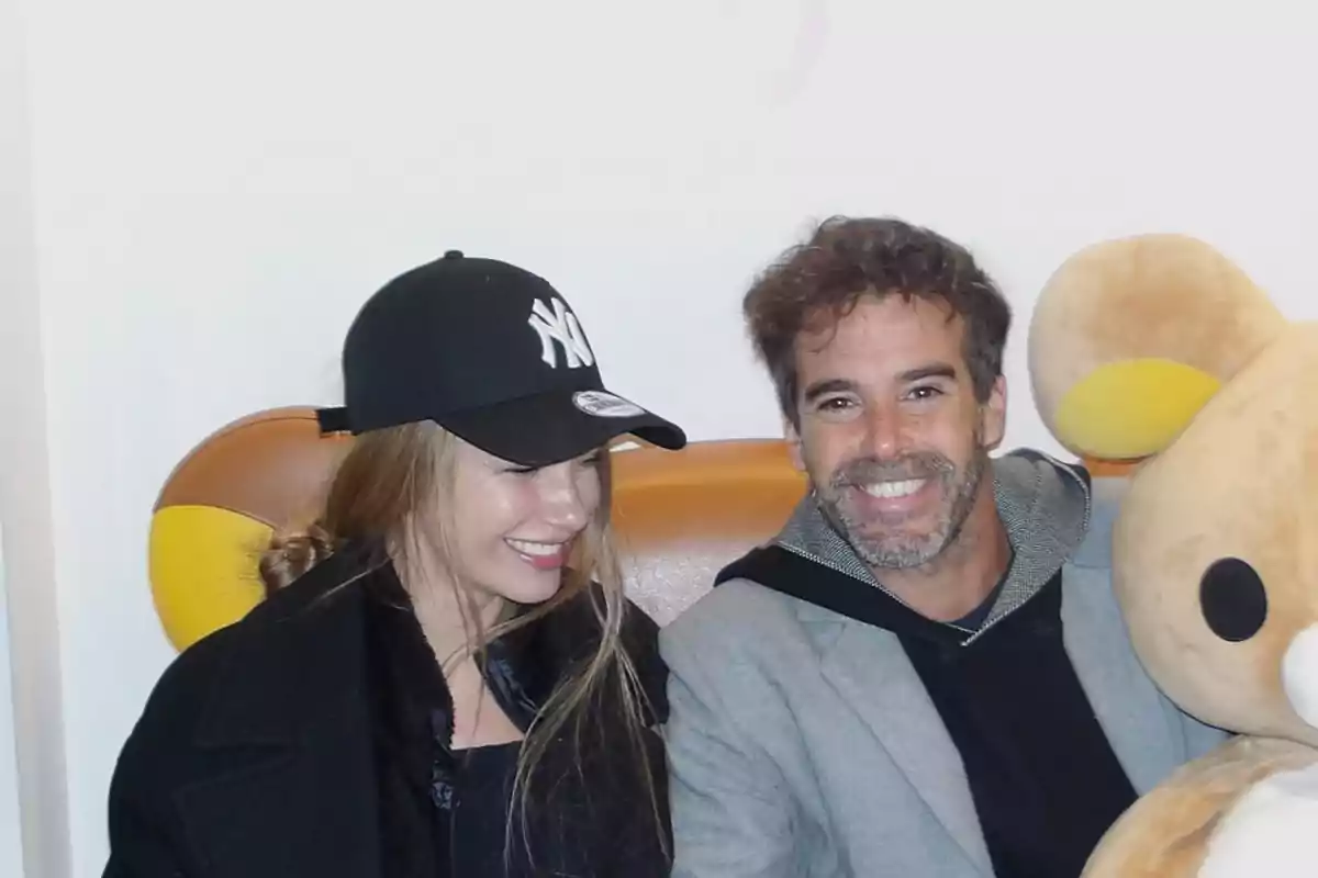 Una pareja sonriente sentada en un sofá junto a un gran peluche. Una pareja sonriente sentada en un sofá junto a un gran peluche.