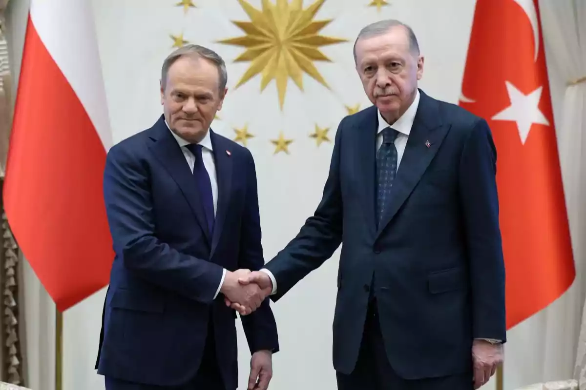 Two men in dark suits shake hands in front of flags and a golden emblem on the wall.