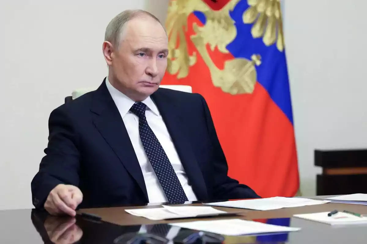 Man in a dark suit sitting at a desk with papers and pens, in the background a flag with red, blue, and gold colors Man in a dark suit sitting at a desk with papers and pens, in the background a flag with red, blue, and gold colors