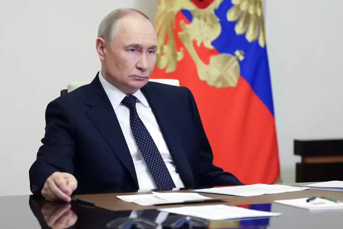 Man in a dark suit sitting at a desk with papers and pens, in the background a flag with red, blue, and gold colors