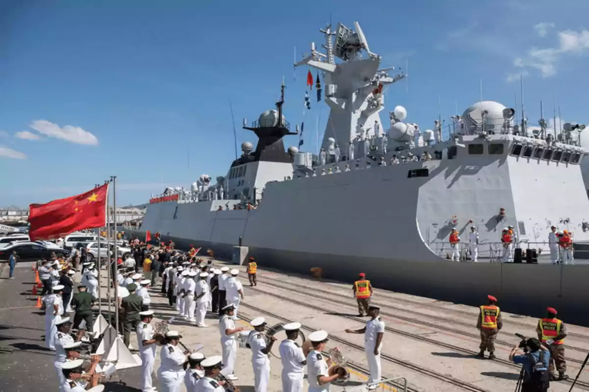 Un buque de guerra atracado en un puerto con marineros alineados en la cubierta y una banda militar tocando en el muelle mientras una bandera roja ondea en el viento.