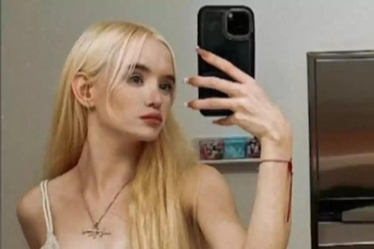 A person with long blonde hair takes a selfie in front of a mirror using a mobile phone.