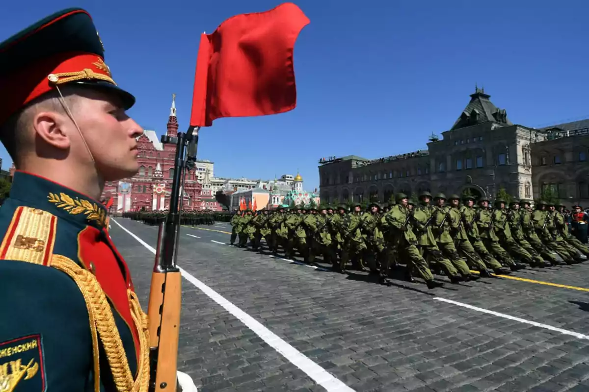 Un soldado con uniforme ceremonial sostiene una bandera roja mientras un grupo de soldados marcha en formación en una plaza con edificios históricos al fondo. Un soldado con uniforme ceremonial sostiene una bandera roja mientras un grupo de soldados marcha en formación en una plaza con edificios históricos al fondo.