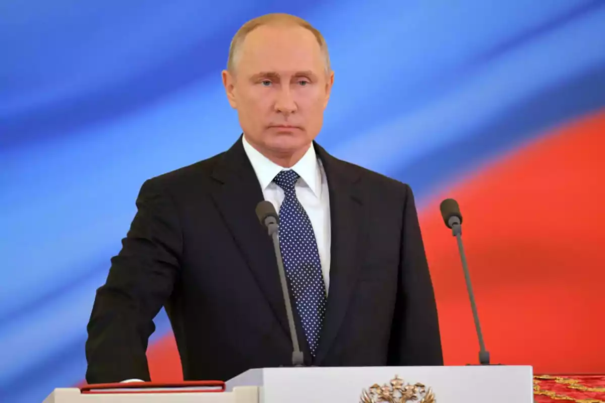 A man in a dark suit and polka dot tie is standing in front of a podium with microphones, against a blue and red background. A man in a dark suit and polka dot tie is standing in front of a podium with microphones, against a blue and red background.