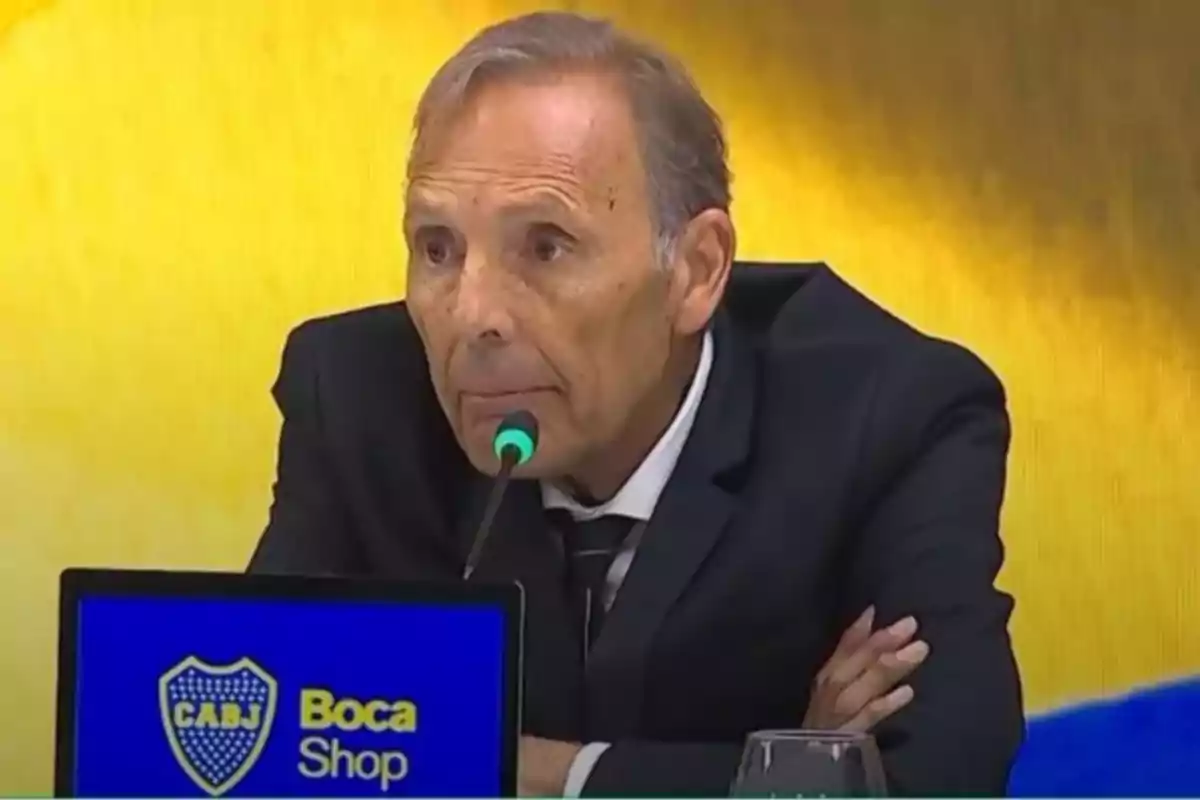 Hombre mayor de traje hablando en una conferencia de prensa con un micrófono frente a una pantalla que muestra el logo de Boca Juniors y la palabra Shop Hombre mayor de traje hablando en una conferencia de prensa con un micrófono frente a una pantalla que muestra el logo de Boca Juniors y la palabra Shop