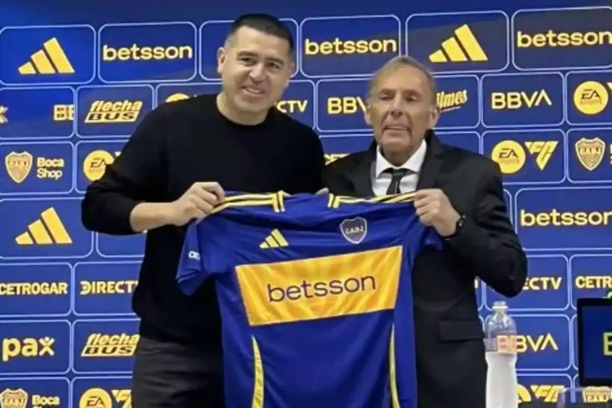 Dos hombres posan sonrientes sosteniendo una camiseta de Boca Juniors frente a un fondo con logotipos de patrocinadores del club