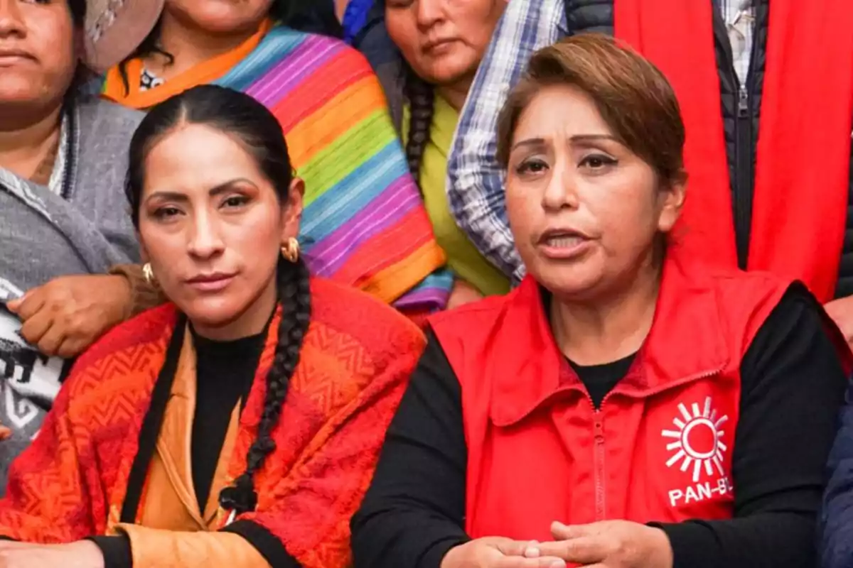 Dos mujeres sentadas al frente de un grupo, una con chal rojo y trenza, la otra con chaleco rojo con el logo de PAN-BOL, ambas con expresión seria Dos mujeres sentadas al frente de un grupo, una con chal rojo y trenza, la otra con chaleco rojo con el logo de PAN-BOL, ambas con expresión seria