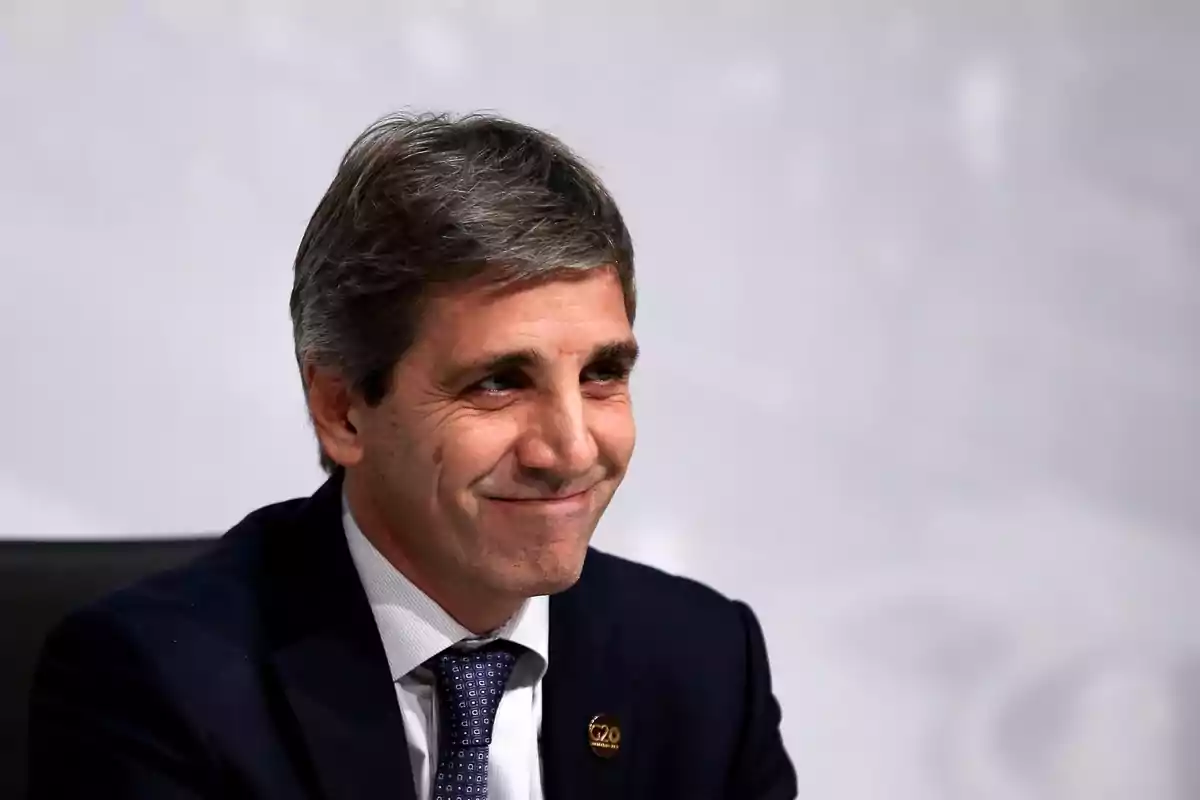 Un hombre con traje oscuro y corbata sonríe mientras está sentado frente a un fondo gris.