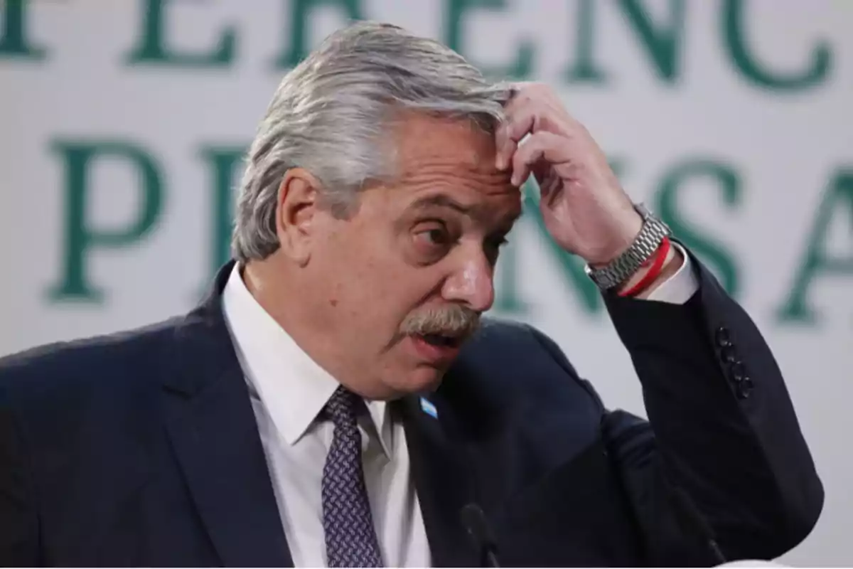 Hombre de cabello canoso y bigote, vestido con traje y corbata, se toca la cabeza con expresión pensativa frente a un micrófono.