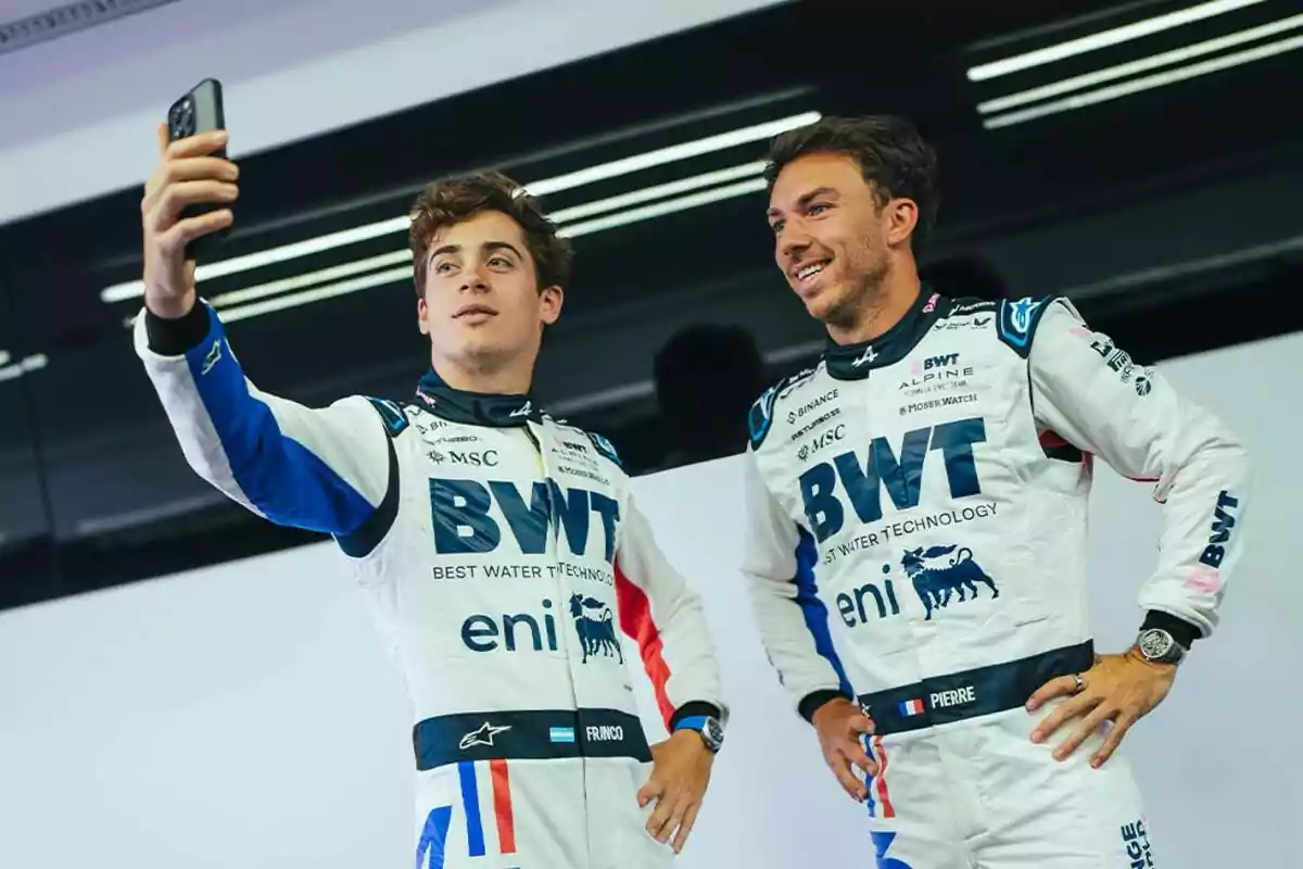 Two drivers in white racing suits with blue details pose for a selfie inside a motorsport garage.