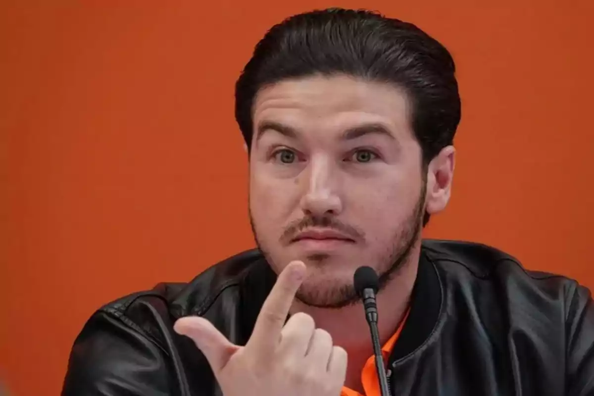 Man in a black jacket and orange jersey speaking into a microphone in front of an orange background.