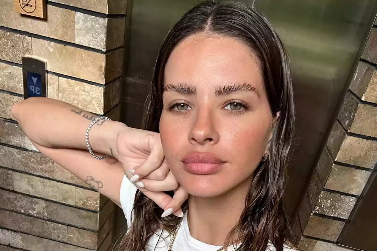 Mujer con cabello mojado y camiseta blanca posando frente a una pared de ladrillos.