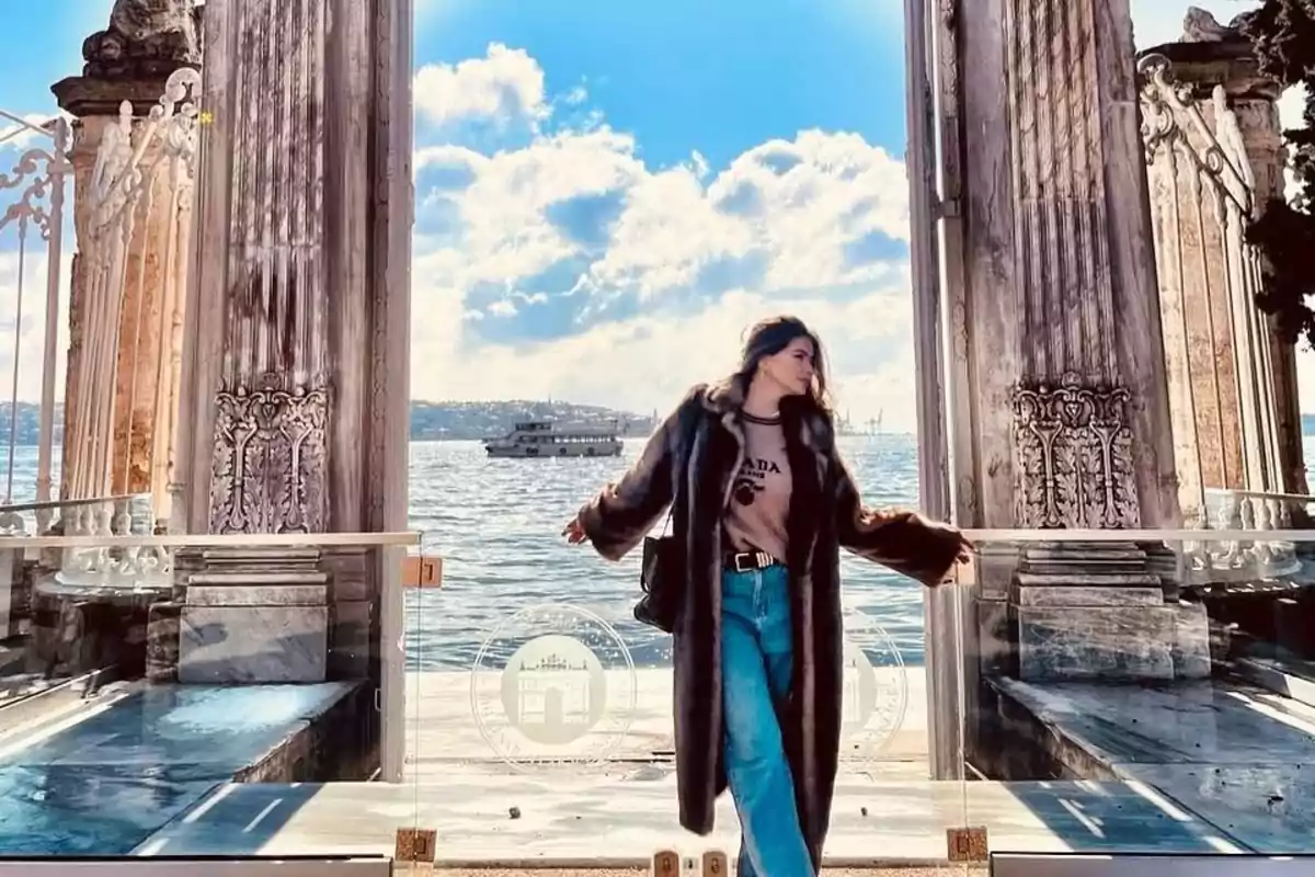 Woman wearing a long coat and jeans posing in front of an old door overlooking the sea with a ship in the background on a sunny day with clouds Woman wearing a long coat and jeans posing in front of an old door overlooking the sea with a ship in the background on a sunny day with clouds