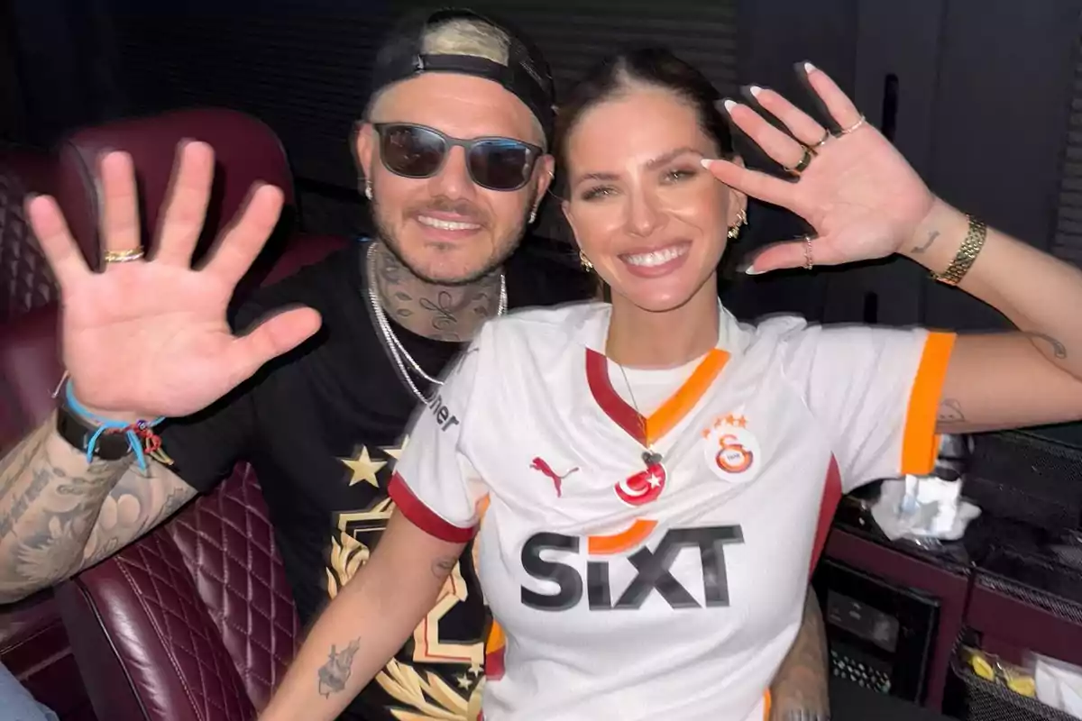 A smiling couple sitting on red leather seats, both raising their hands and looking at the camera; the woman is wearing a white Galatasaray soccer jersey and the man is wearing sunglasses and a black cap. A smiling couple sitting on red leather seats, both raising their hands and looking at the camera; the woman is wearing a white Galatasaray soccer jersey and the man is wearing sunglasses and a black cap.