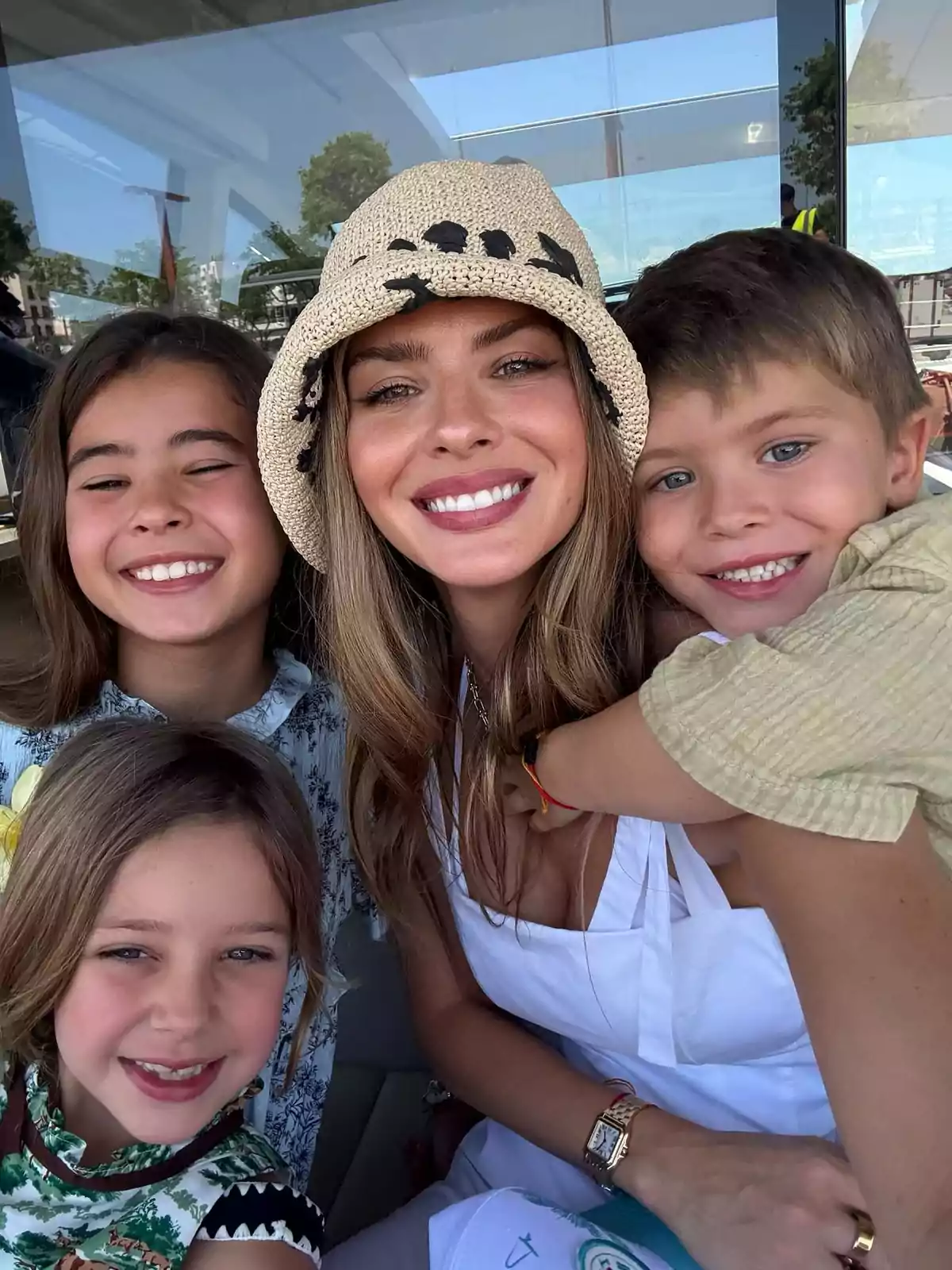 Una mujer sonriente con sombrero claro posa junto a tres niños que también sonríen, todos en un ambiente al aire libre y soleado. Una mujer sonriente con sombrero claro posa junto a tres niños que también sonríen, todos en un ambiente al aire libre y soleado.