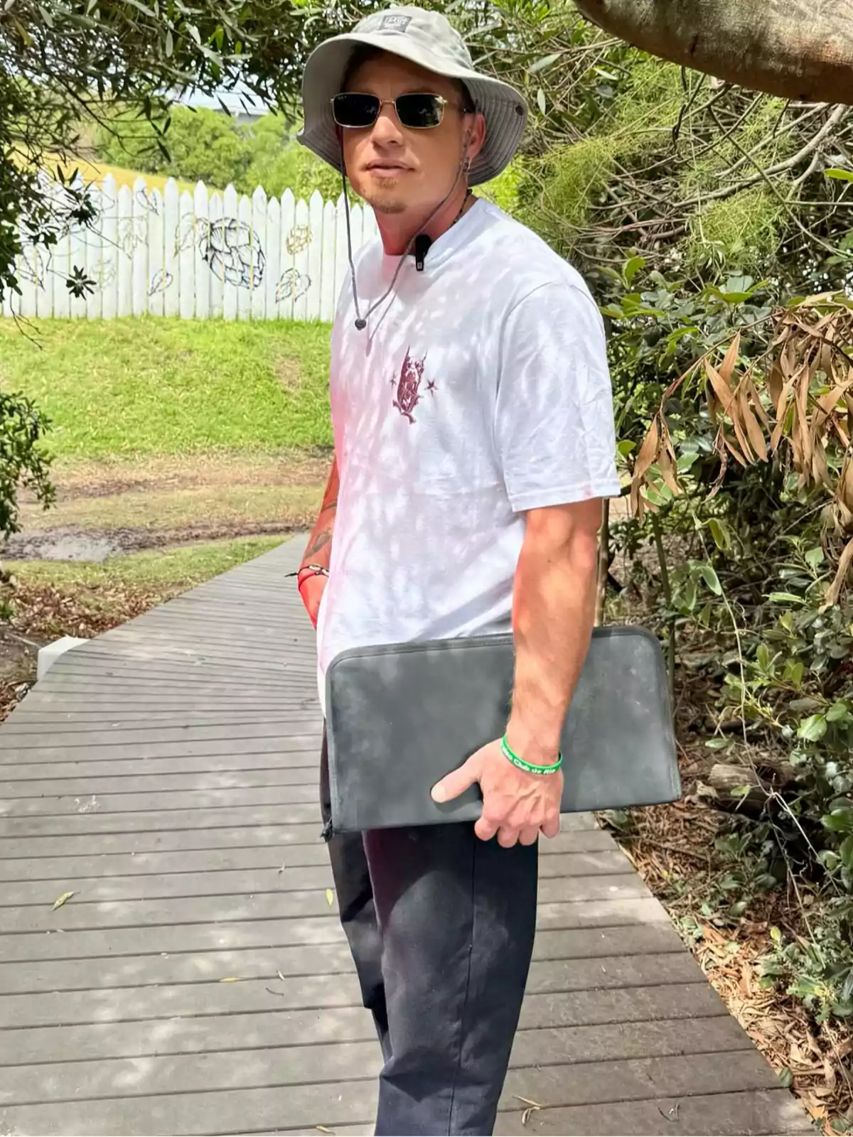 Un hombre con sombrero y gafas de sol sostiene una funda negra mientras camina por un sendero de madera rodeado de vegetación. Un hombre con sombrero y gafas de sol sostiene una funda negra mientras camina por un sendero de madera rodeado de vegetación.