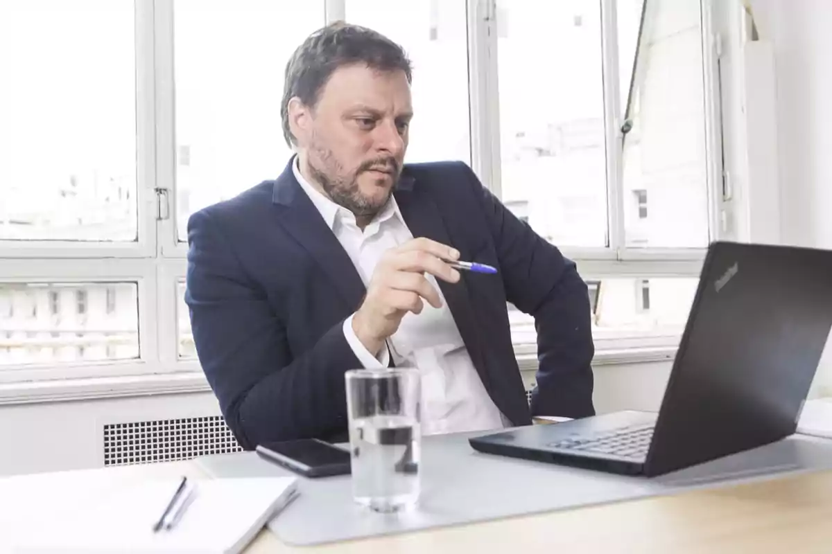 A man in a dark suit sitting at a desk in front of a laptop holding a pen with a thoughtful expression.