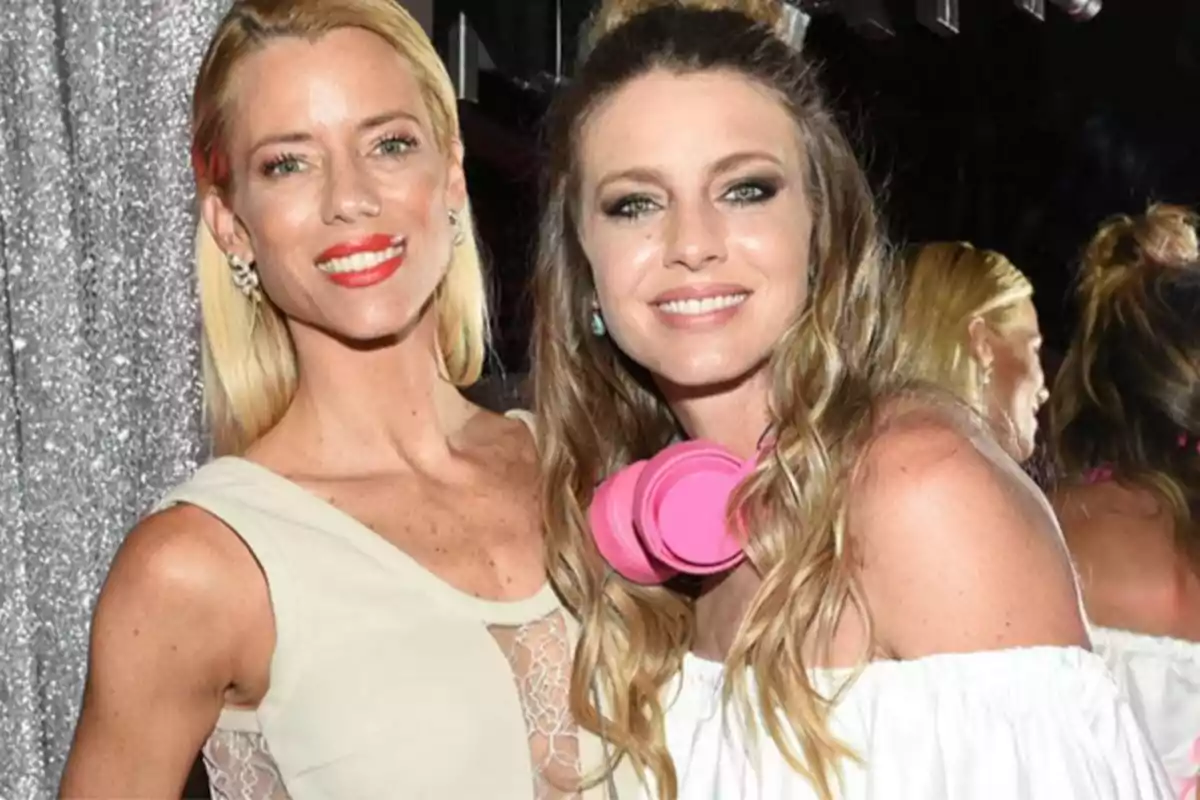 Two women smile at a party, one wearing pink headphones around her neck and the other wearing a light-colored dress.