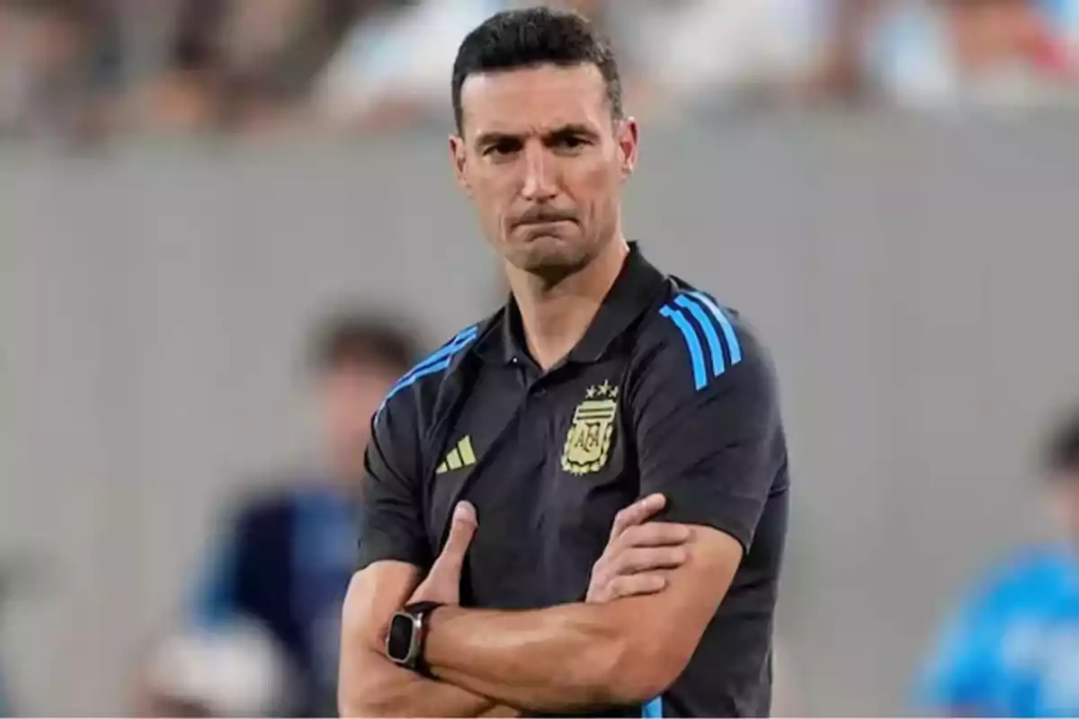 A man in a black sports jersey with blue details crosses his arms while observing something with a serious expression. A man in a black sports jersey with blue details crosses his arms while observing something with a serious expression.