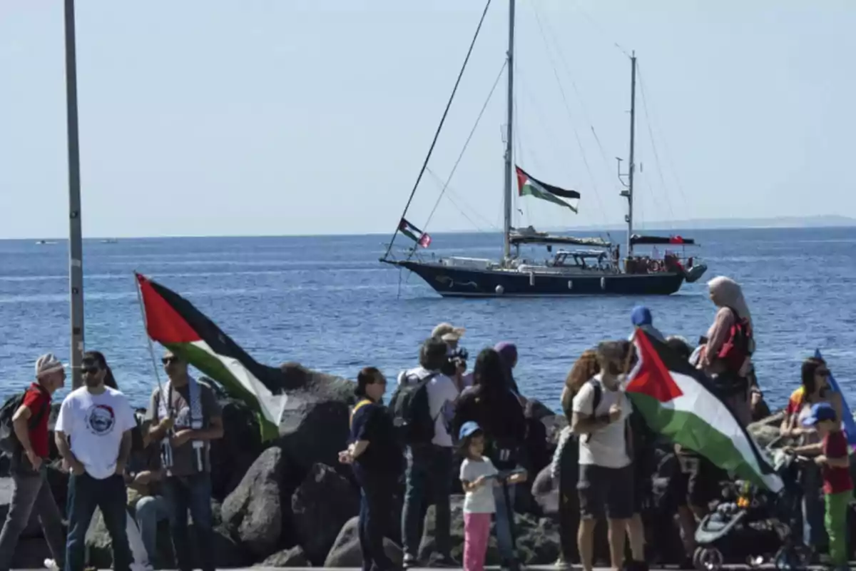 People gathered by the sea holding Palestinian flags while a sailboat with the same flag sails near the shore People gathered by the sea holding Palestinian flags while a sailboat with the same flag sails near the shore