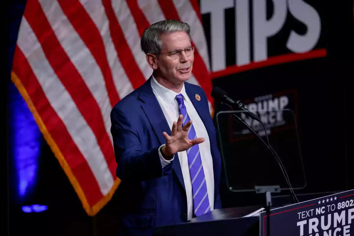Gray-haired man with glasses speaking at a podium with a microphone, wearing a blue suit and a purple tie; in the background, there is a United States flag and part of a campaign poster. Gray-haired man with glasses speaking at a podium with a microphone, wearing a blue suit and a purple tie; in the background, there is a United States flag and part of a campaign poster.