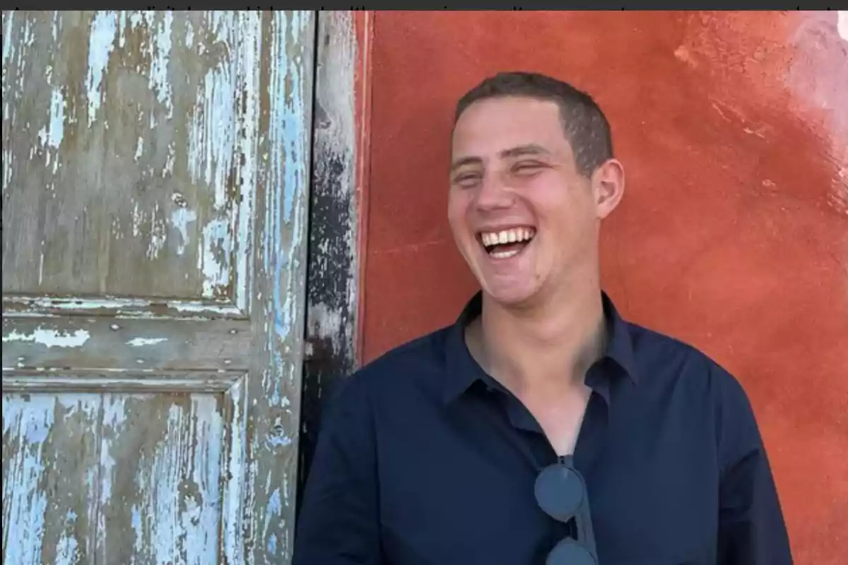 Un hombre sonriente con camisa oscura y gafas colgando de su cuello está de pie junto a una puerta de madera desgastada y una pared de color terracota.