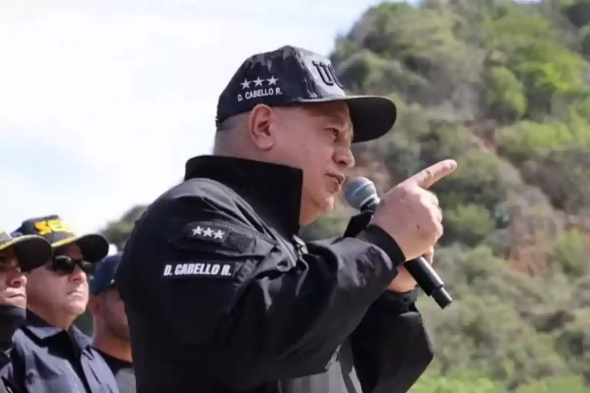 Hombre con gorra y uniforme negro hablando por un micrófono al aire libre con fondo de vegetación.