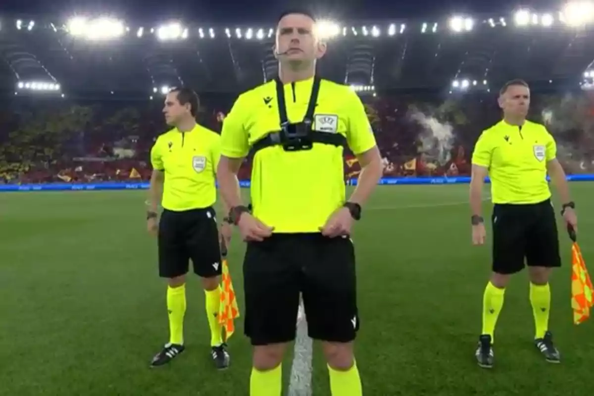 Three soccer referees in yellow uniforms pose on the field before a match in a brightly lit stadium full of fans. Three soccer referees in yellow uniforms pose on the field before a match in a brightly lit stadium full of fans.