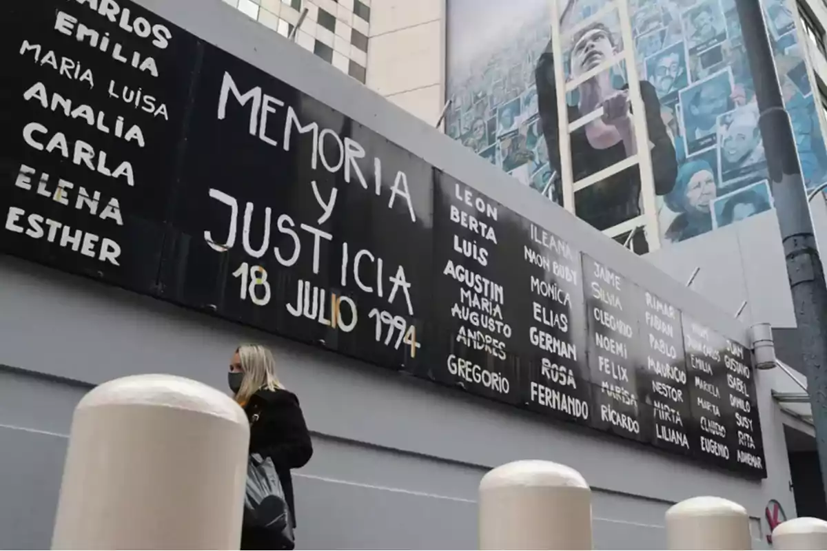 Una mujer con mascarilla camina frente a un mural que dice "Memoria y Justicia 18 Julio 1994" y tiene una lista de nombres.