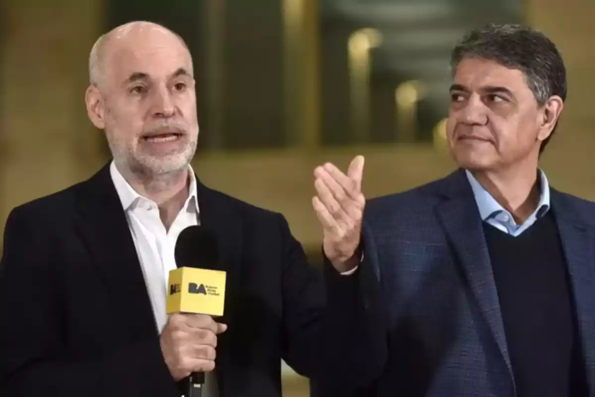 Dos hombres de traje, uno de ellos sosteniendo un micrófono amarillo con el logo "BA", están hablando en un entorno interior.