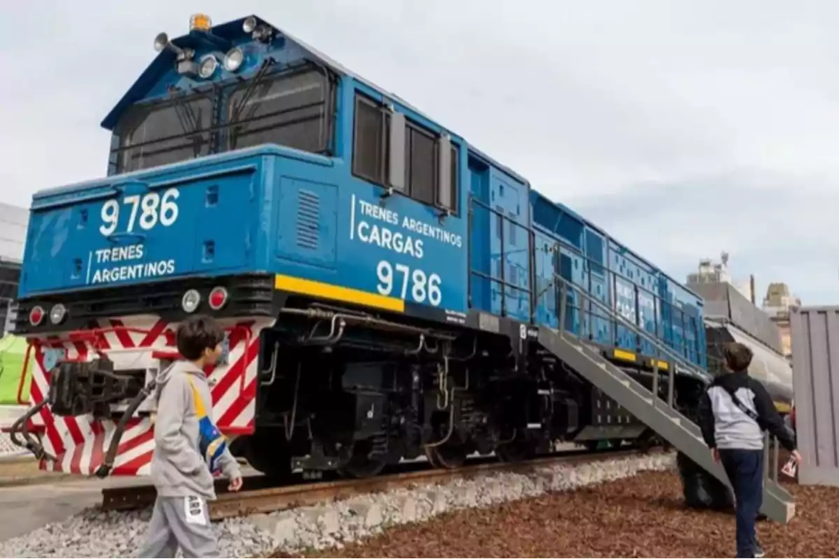 Un tren de carga azul con el número 9786 y la inscripción "Trenes Argentinos Cargas" está estacionado en una vía, mientras dos niños observan y caminan cerca de él.