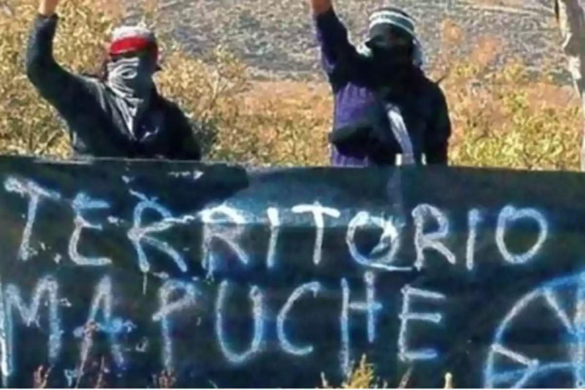 Dos personas con el rostro cubierto levantan el puño detrás de una pancarta que dice "TERRITORIO MAPUCHE".