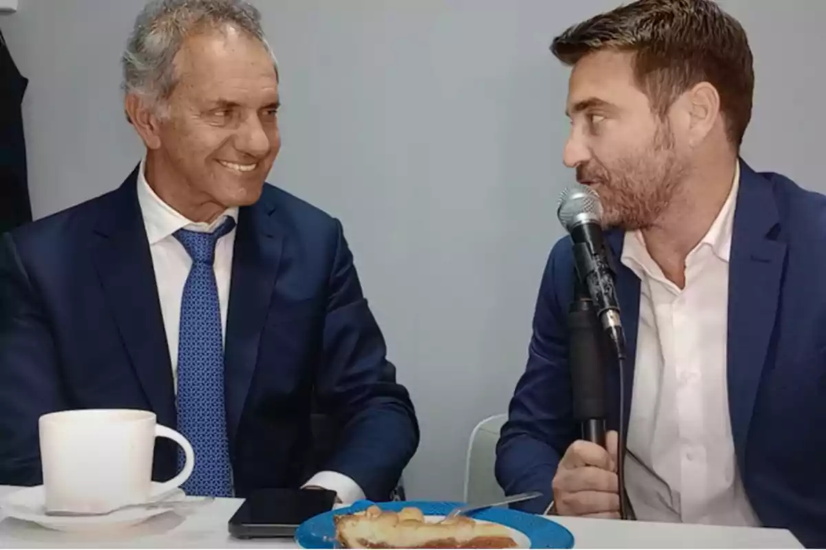 Dos hombres en trajes formales conversan mientras uno de ellos sostiene un micrófono, con una taza de café y un plato con pastel sobre la mesa.