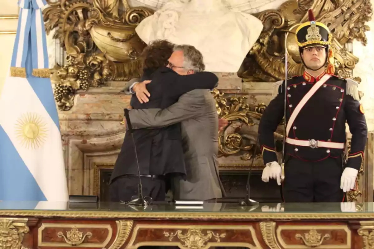 Dos personas se abrazan frente a una bandera argentina y un guardia uniformado en un entorno decorado con ornamentos dorados y una escultura de mármol.