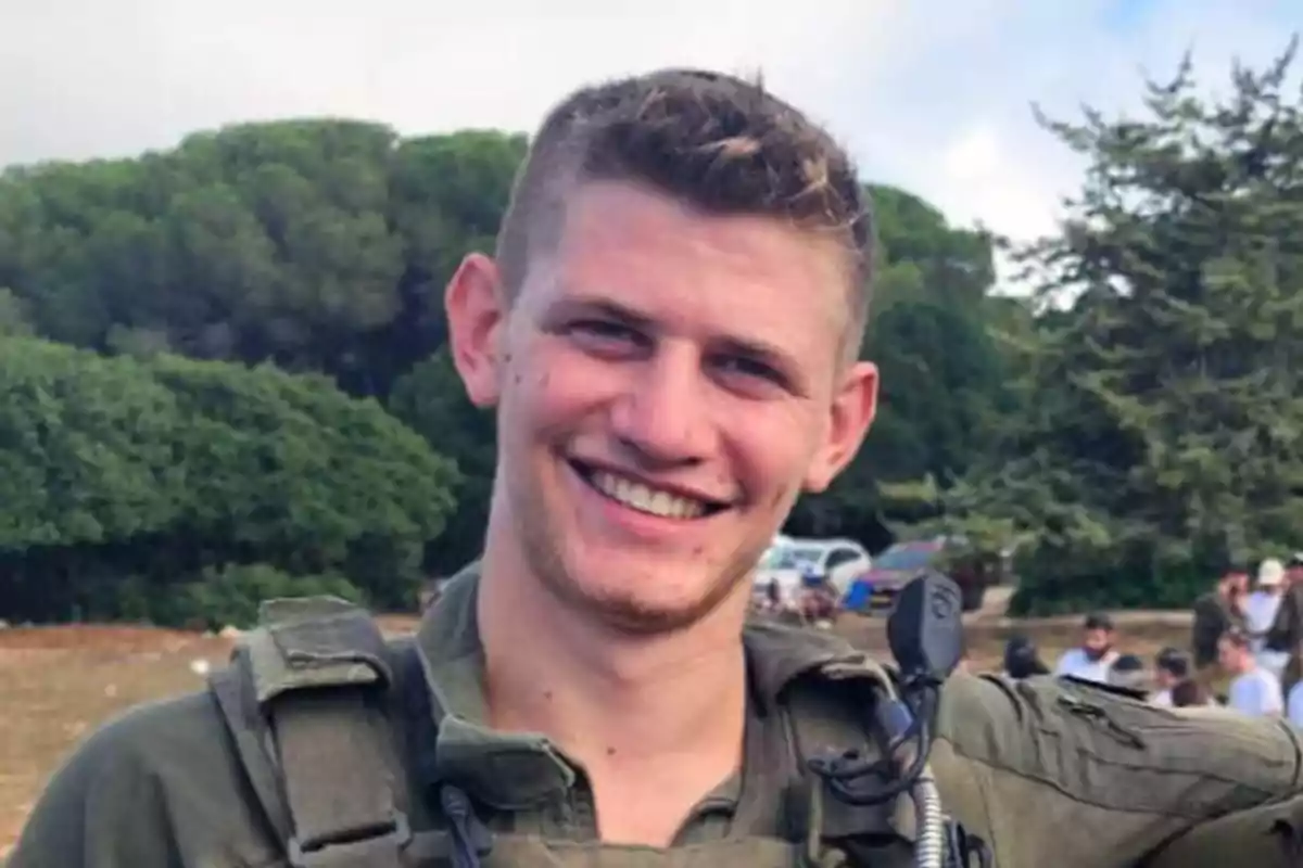 Un hombre joven con uniforme militar sonriendo al aire libre con árboles y personas en el fondo.