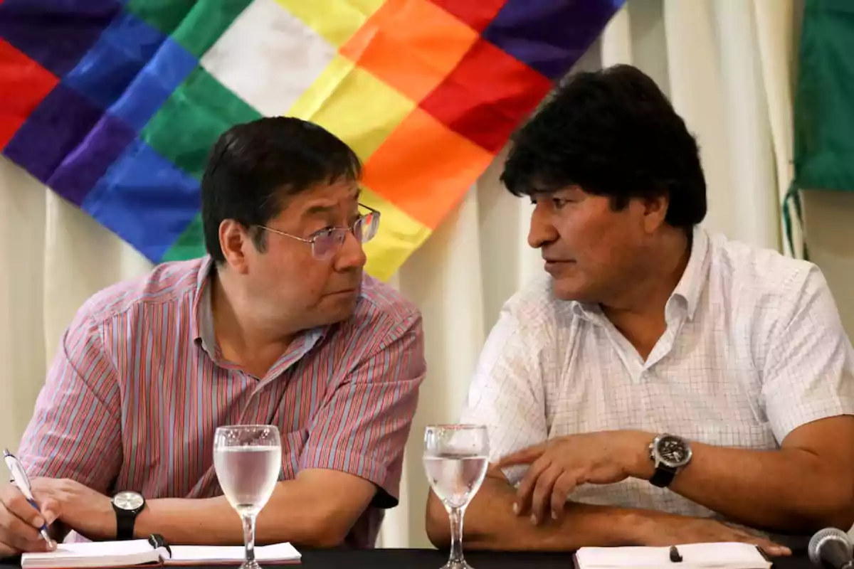 Dos hombres conversan en una mesa con copas de agua y cuadernos, con una bandera colorida en el fondo.