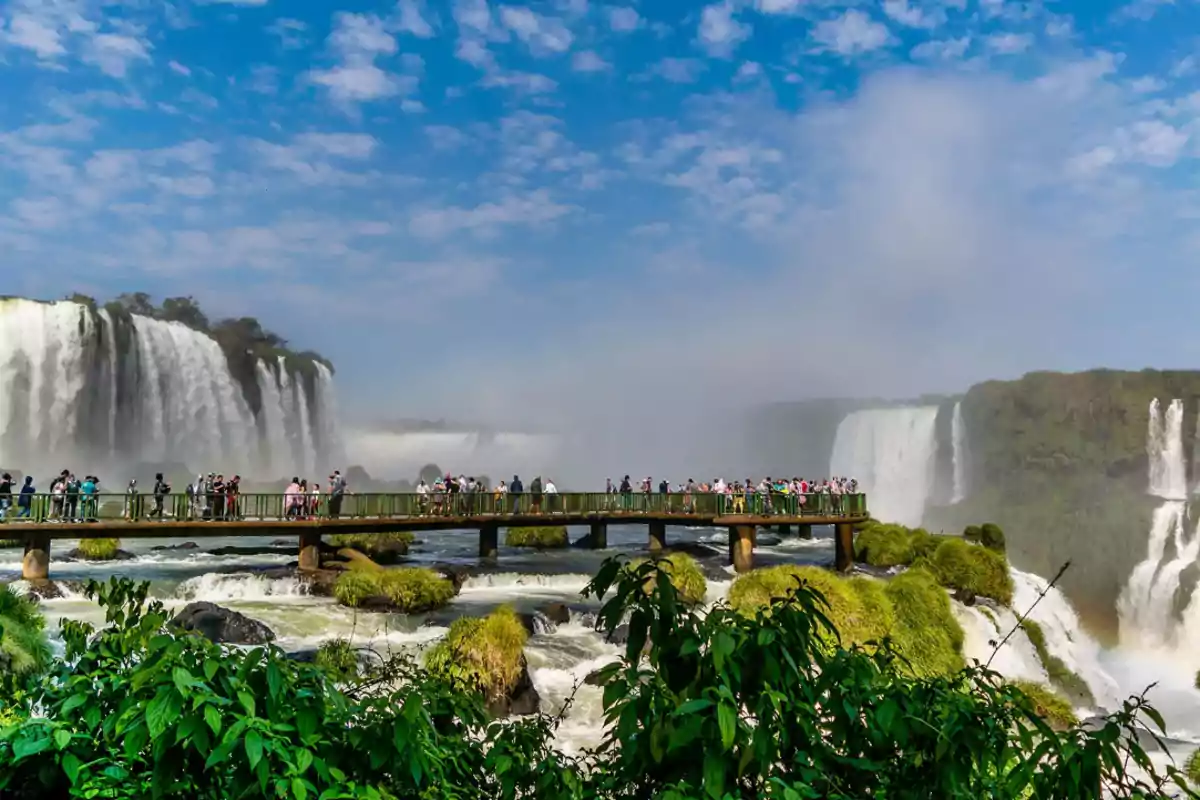 Personas caminando por una pasarela frente a las impresionantes cataratas rodeadas de vegetación exuberante bajo un cielo azul.