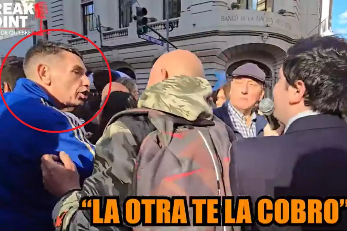 Un grupo de personas discute en la calle frente a un edificio con la inscripción "Banco de la Nación Argentina" mientras un hombre sostiene un micrófono y hay un texto en la parte inferior que dice "La otra te la cobro".