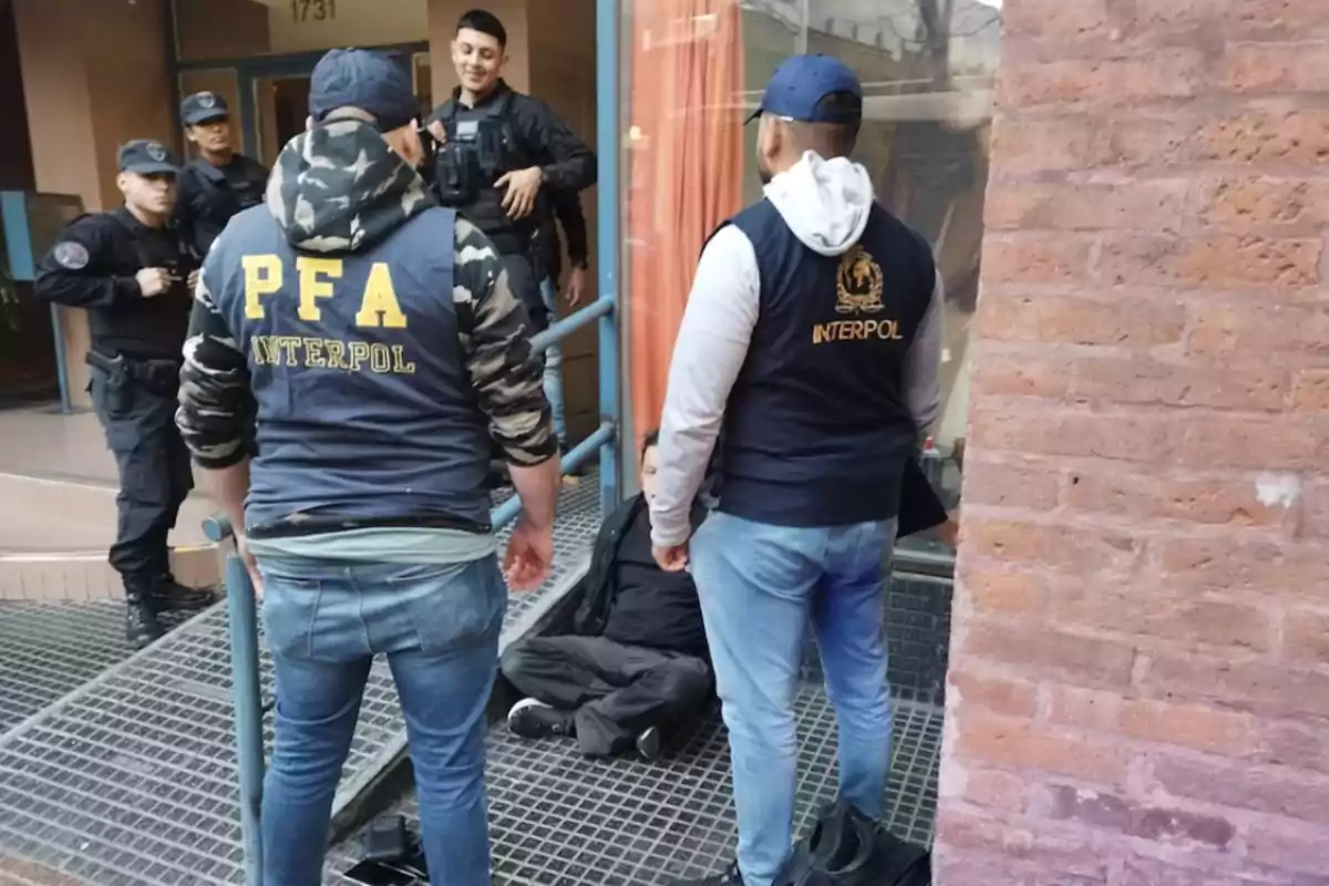 Un grupo de agentes de la PFA e Interpol detiene a una persona en la entrada de un edificio mientras otros oficiales observan.