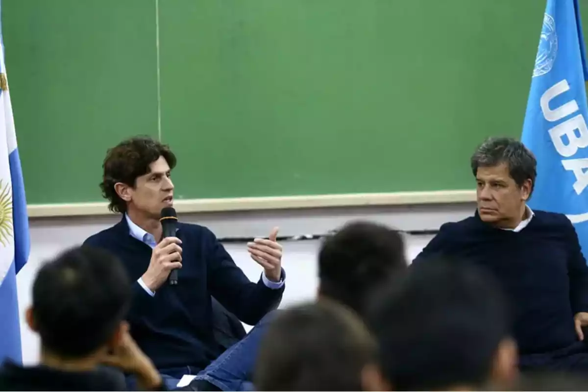 Dos personas sentadas en un aula frente a una pizarra verde, una de ellas sostiene un micrófono mientras habla, y hay una bandera de la UBA visible al fondo.