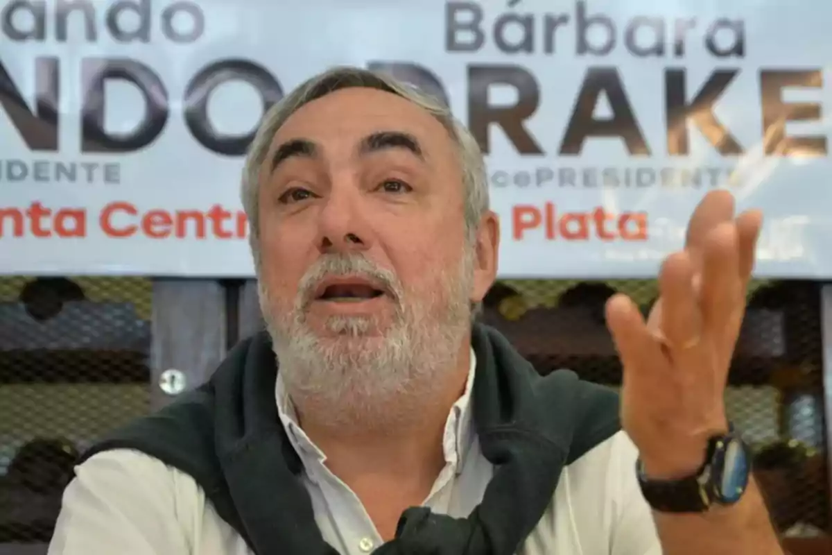 Un hombre con barba y cabello canoso gesticula mientras está sentado frente a un cartel que tiene texto parcialmente visible.