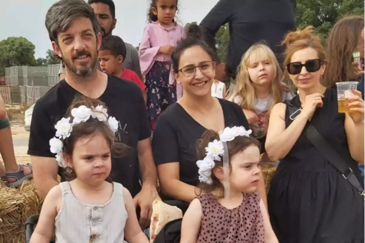 Un grupo de personas, incluyendo adultos y niños, posan al aire libre en un ambiente festivo; dos niñas al frente llevan diademas con flores blancas.