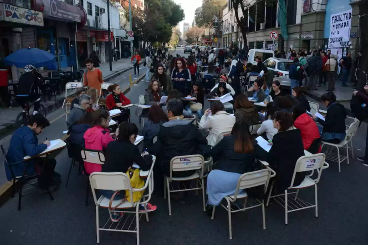 Un grupo de personas se encuentra sentado en sillas y escritorios en medio de una calle, participando en una actividad al aire libre, mientras otras personas caminan alrededor y se observan edificios y negocios en el fondo.