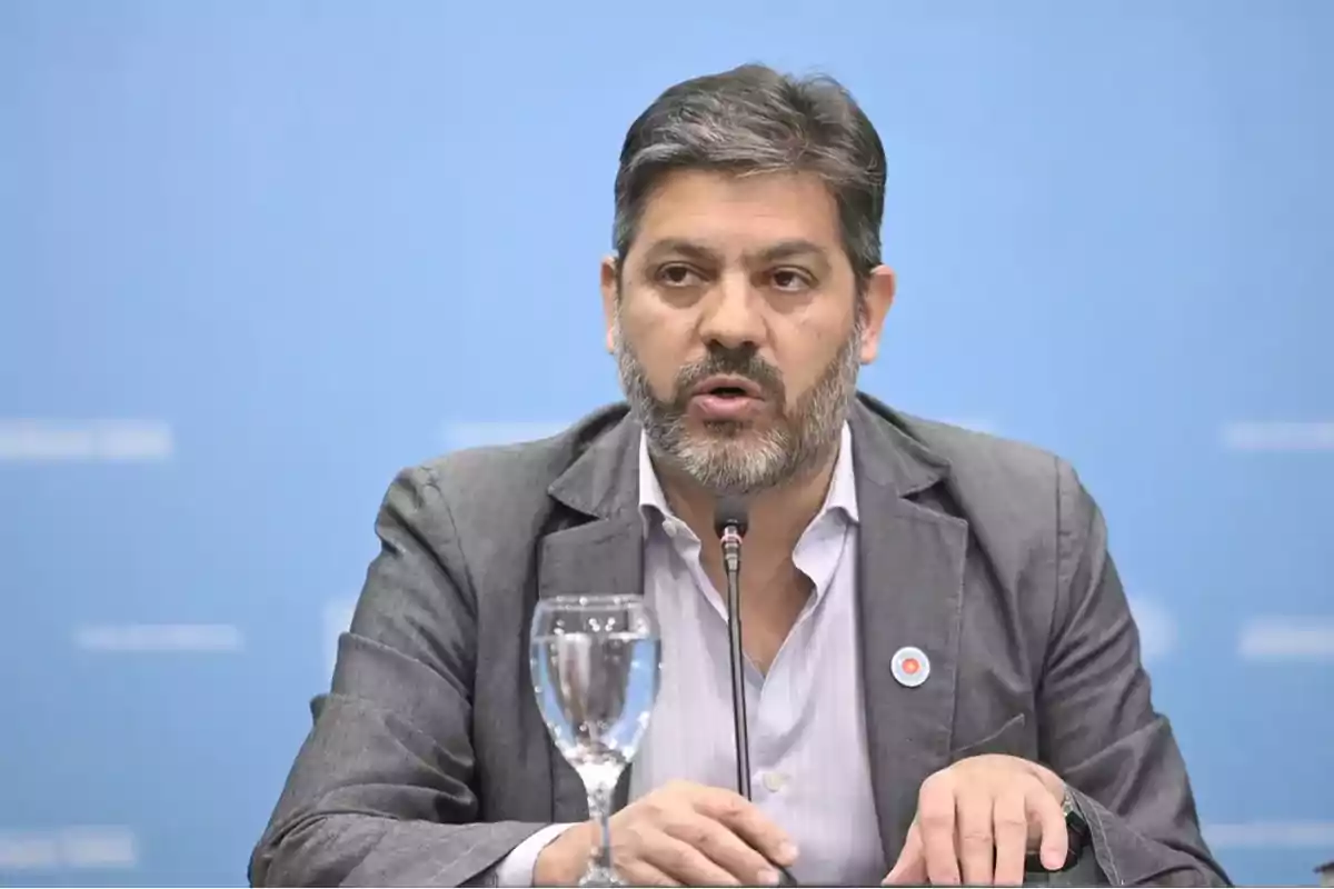 Hombre con barba hablando en una conferencia de prensa con un micrófono y un vaso de agua frente a él.