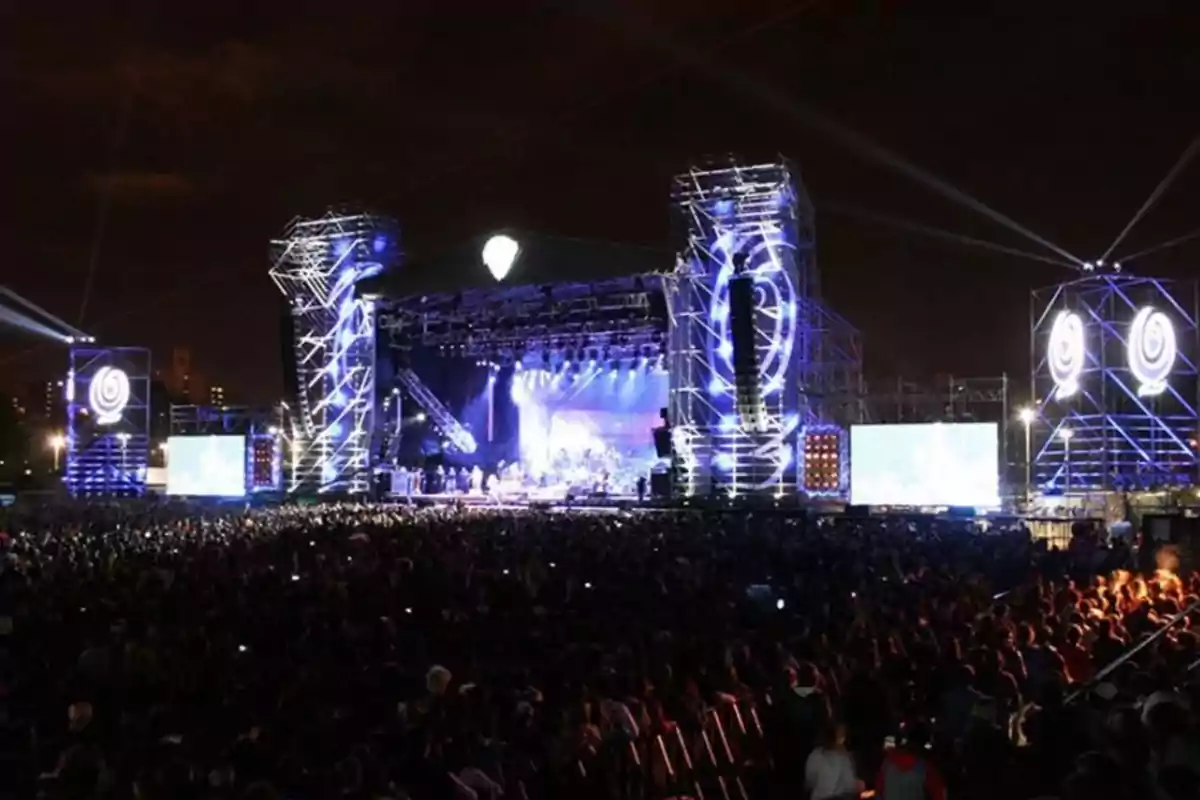 Una multitud disfruta de un concierto nocturno en un gran escenario iluminado con luces azules y pantallas gigantes.