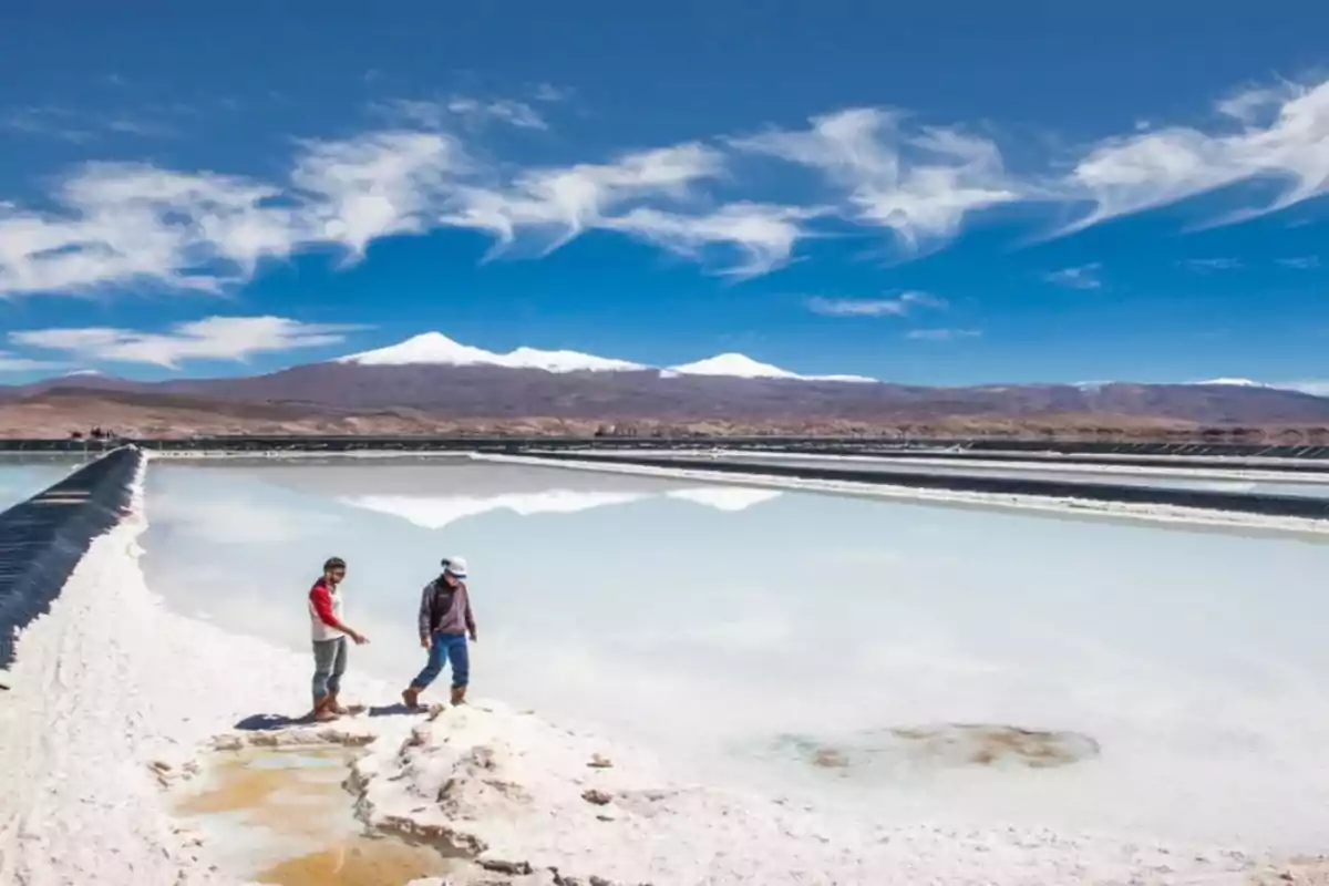 Dos personas caminan sobre una salina blanca junto a un cuerpo de agua reflectante con montañas nevadas al fondo bajo un cielo azul con nubes dispersas.