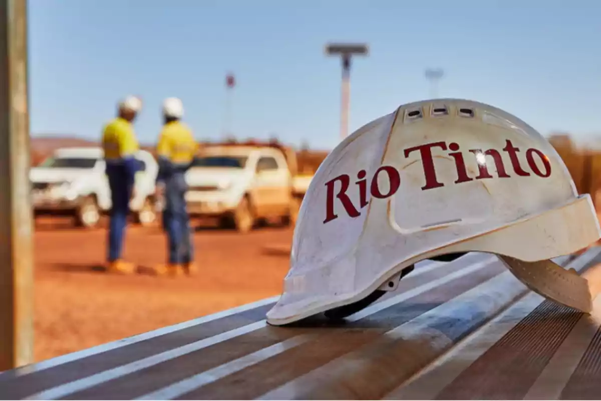 Casco de seguridad con el logo de Rio Tinto en primer plano y dos trabajadores con cascos y chalecos en el fondo junto a vehículos en un entorno industrial.