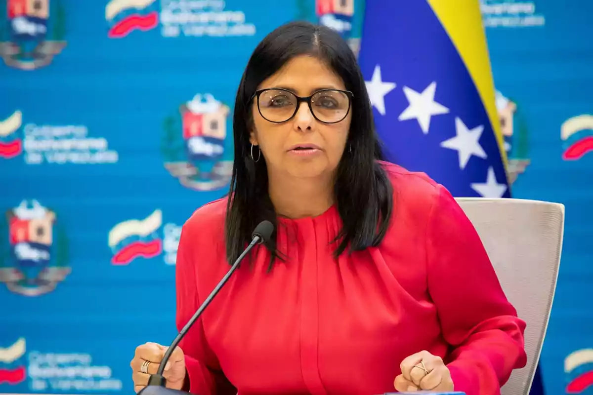 Una persona con gafas y blusa roja hablando frente a un micrófono con un fondo azul que tiene emblemas y una bandera.