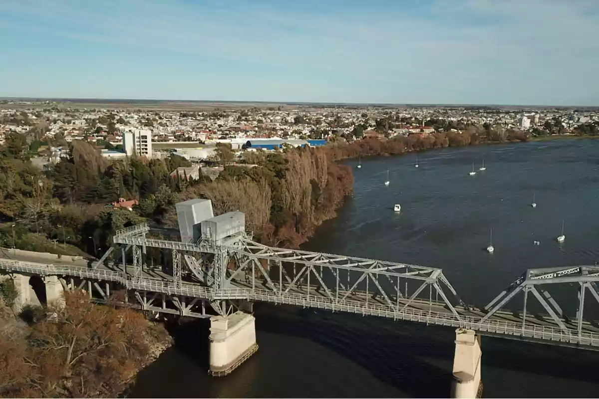 Vista aérea de un puente metálico sobre un río con barcos pequeños y una ciudad al fondo.