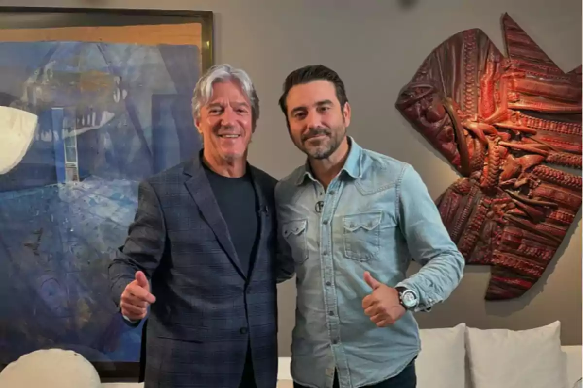 Dos hombres posan juntos sonriendo y levantando el pulgar en una sala con una pintura azul y una escultura de pez en la pared.